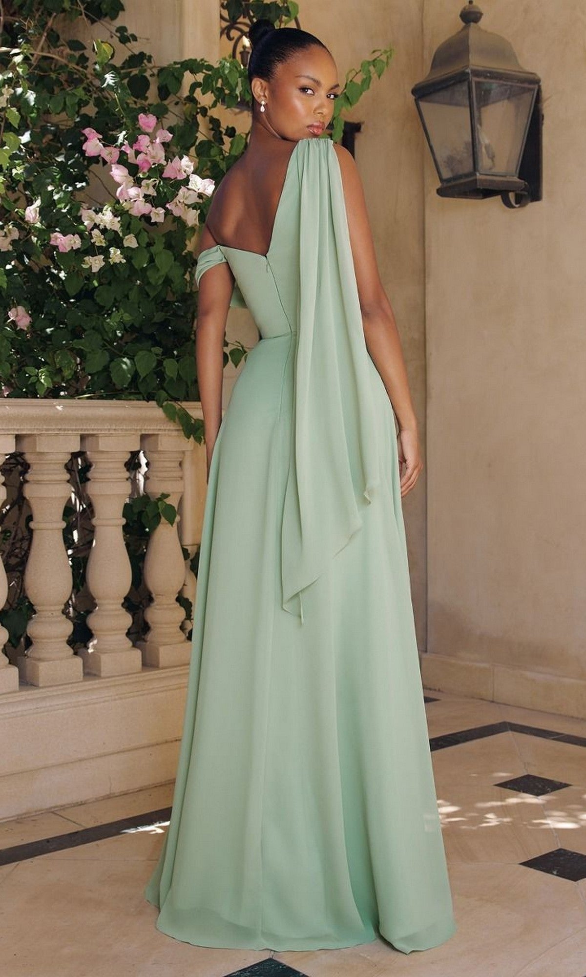 A woman stands on a tiled patio wearing the affordable Asymmetric Long A-Line Prom Dress: Amoris T716, a light green off-shoulder gown with a draped sash, facing away. Elegant plants and a lantern decorate the background.